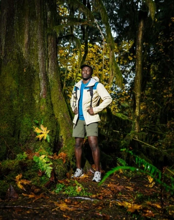 Black man wearing a beige jacket and shorts standing on a mossy forest path by a large tree trunk.