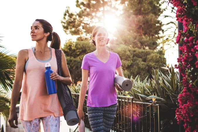 Two women carrying yoga mats and water bottles walking together outdoors near bright pink flowers.