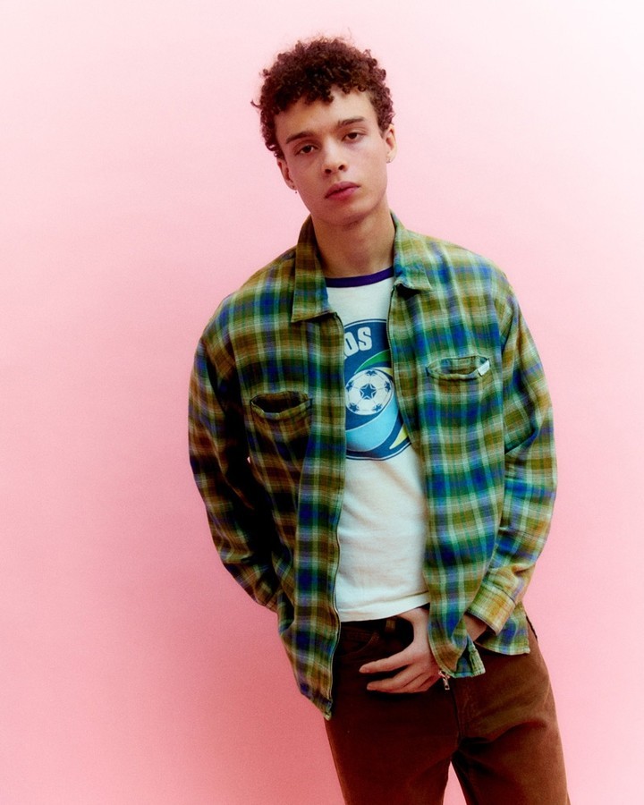 Young adult with curly hair poses in an unbuttoned green and blue plaid shirt over a graphic tee against a solid pink backdrop.