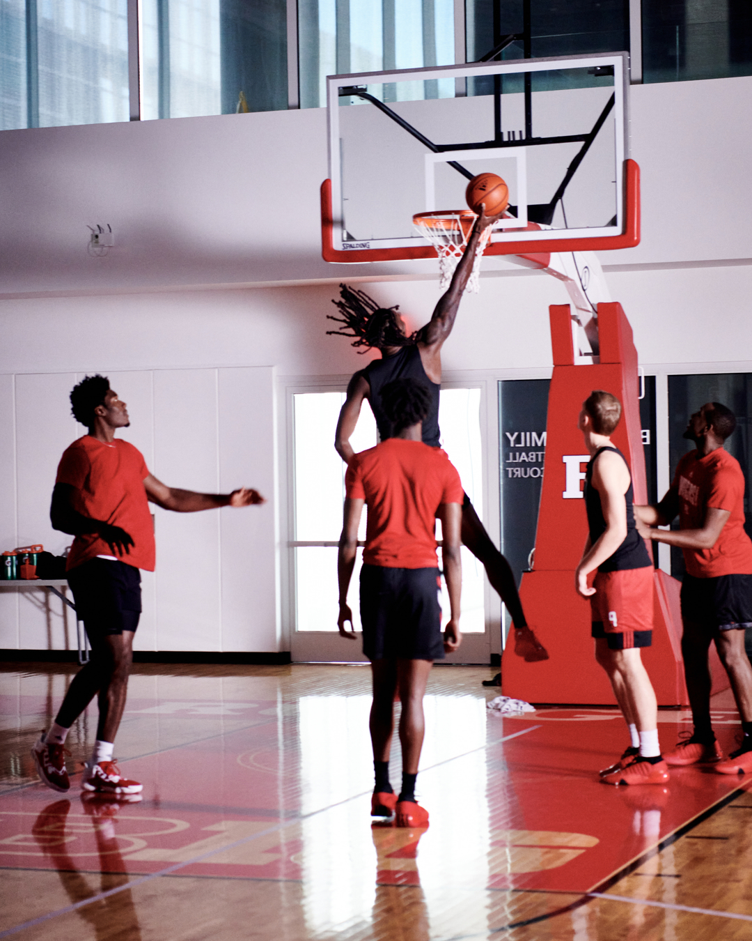 Five basketball players on an indoor court, one leaping toward the hoop with a ball in hand, wearing sports attire and athletic shoes.