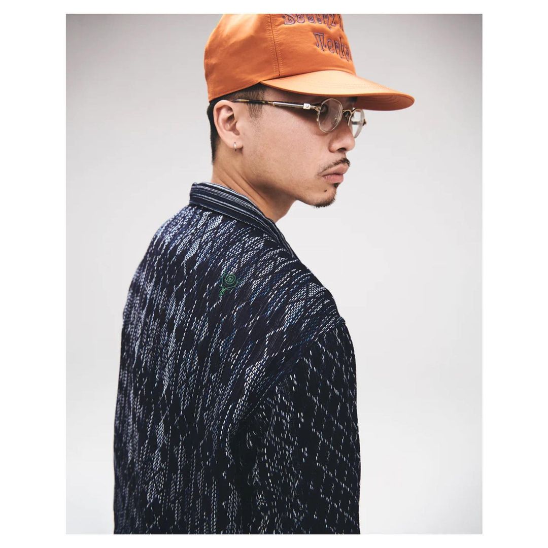 Stylish man in patterned jacket and orange cap, wearing glasses, gazes off to the side against a neutral backdrop in a fashion editorial.
