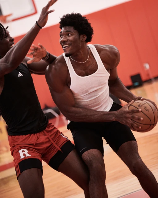 Two athletic men in a gym, one dribbling a basketball, the other defending, showcasing athleticism and sports action in an indoor setting.