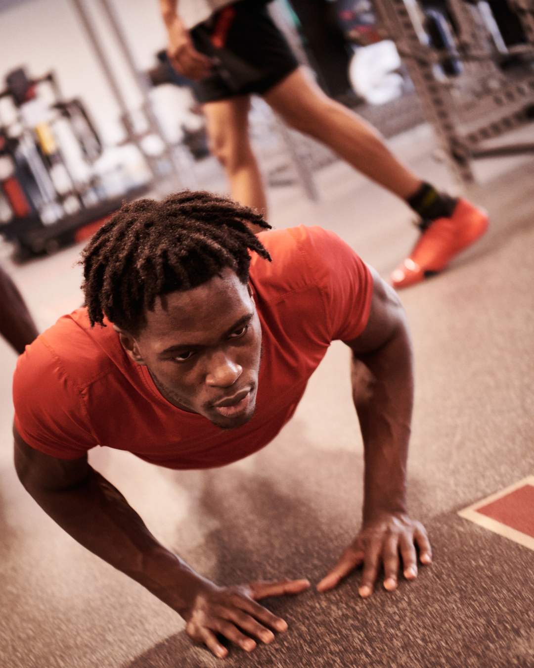 Athletic African American man in red shirt doing push-ups in a gym, with another person walking in background, fitness concept.