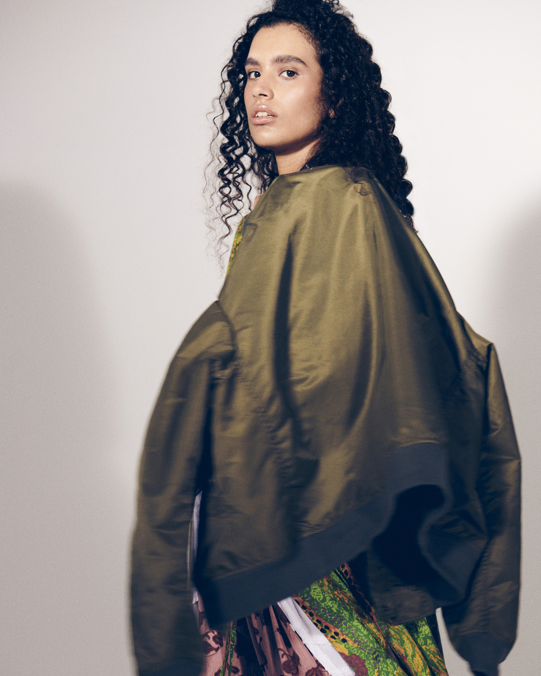 Woman in olive green bomber jacket and patterned skirt posing against a white wall in a fashion-style shot. Studio lighting and curly hair.