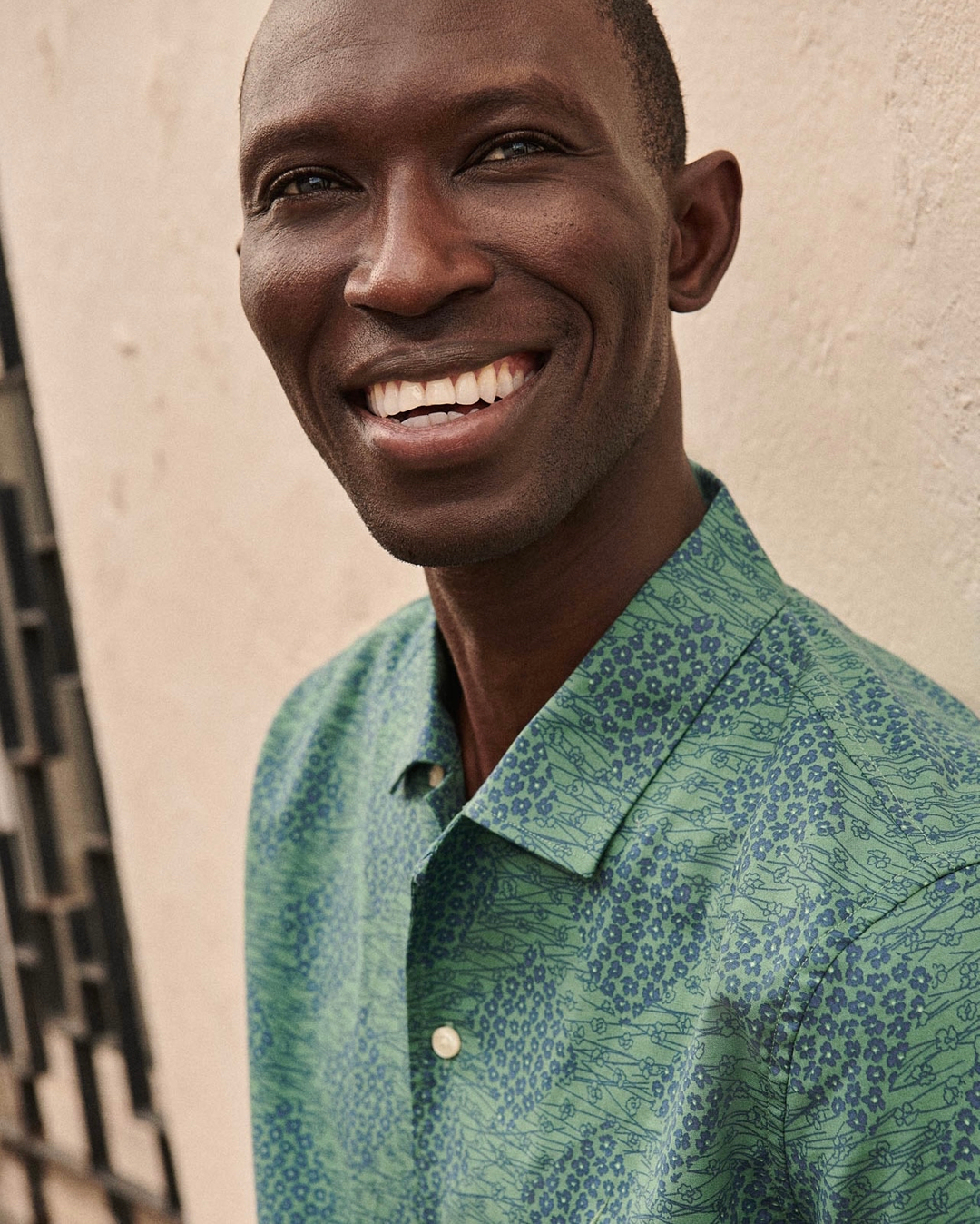 Smiling man with a dark complexion poses in a light green shirt with a floral print next to a tan wall in a medium shot.