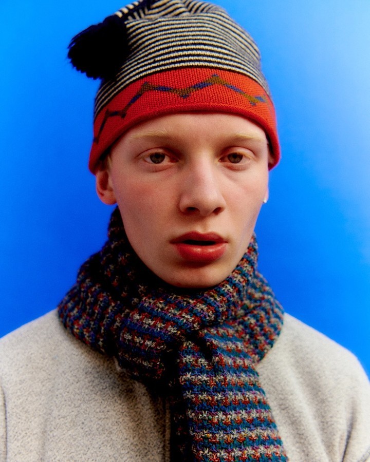 Close-up studio shot of an adult man with fair skin and red hair, wearing a patterned knitted hat and scarf, against a bright blue background.
