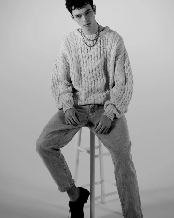 Young man sitting on a white stool wearing a textured knit sweater and jeans against a white seamless background