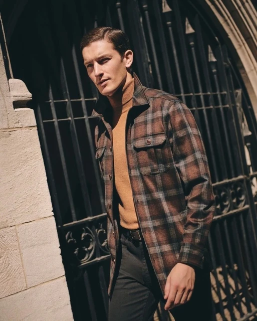 A man in a plaid shirt stands in front of a dark iron gate, wearing a tan turtleneck sweater and dark pants in an urban setting.