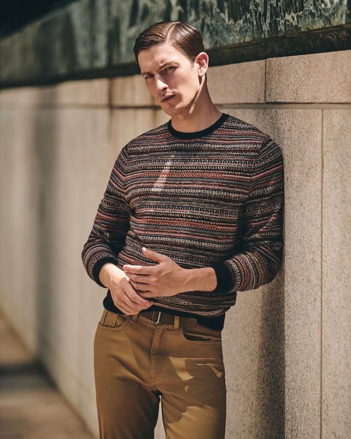Portrait of a young adult male leaning against a light brown wall wearing a patterned sweater and tan trousers with a dark brown belt.