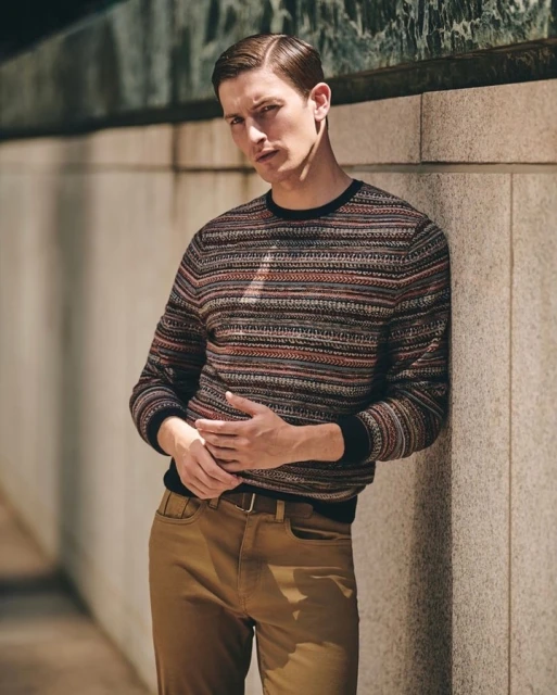 Portrait of a young adult male leaning against a light brown wall wearing a patterned sweater and tan trousers with a dark brown belt.