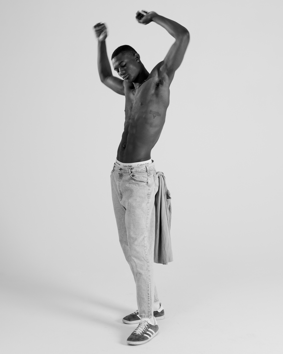 Shirtless black man wearing light wash denim jeans and sneakers with arms raised in studio portrait