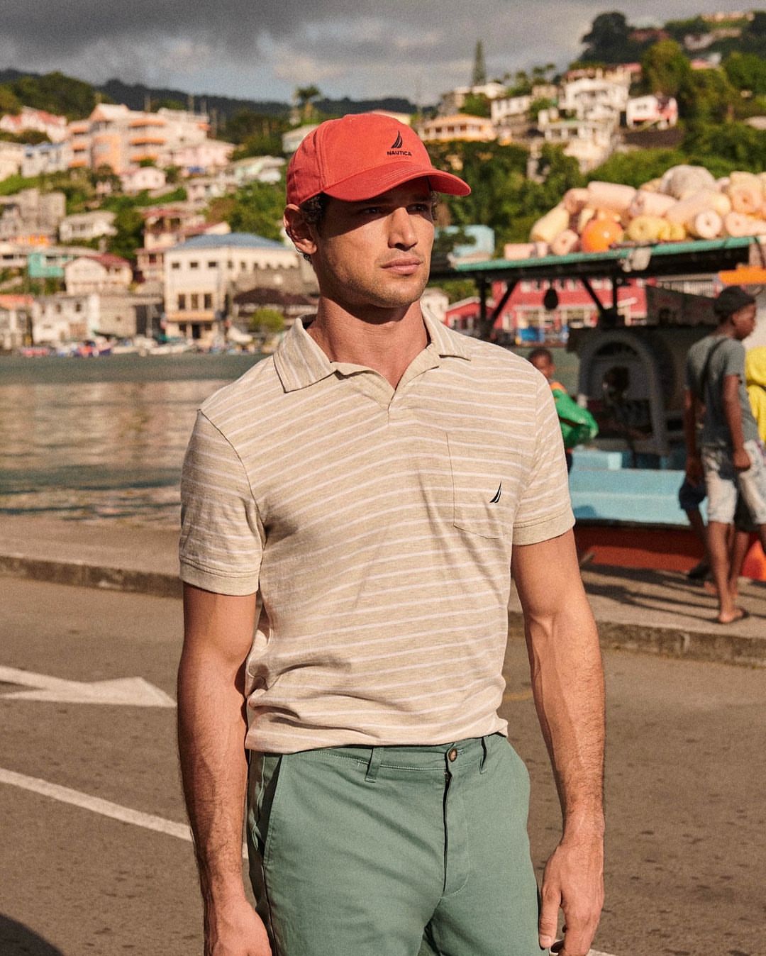 Adult male in a red cap and striped polo shirt standing outside against a background of colorful buildings and vegetation.
