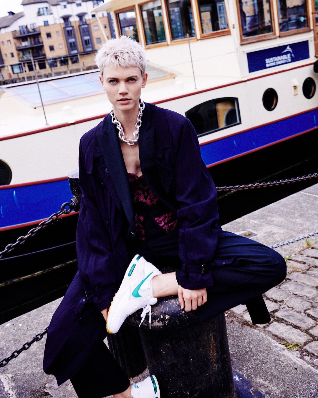 Adult female poses in sportswear and a coat in front of a waterway. She is sitting on a bollard near a boat on a sunny day.