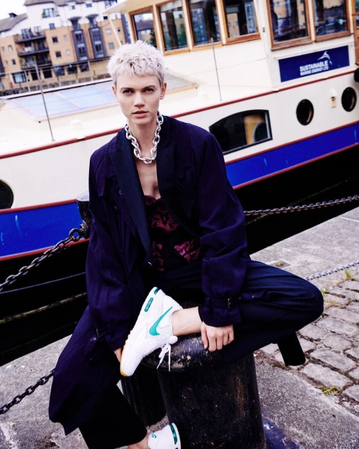 Adult female poses in sportswear and a coat in front of a waterway. She is sitting on a bollard near a boat on a sunny day.