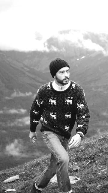 Monochrome outdoor portrait of an adult man wearing a knit hat and patterned sweater walking on a hillside with mountains in the background.