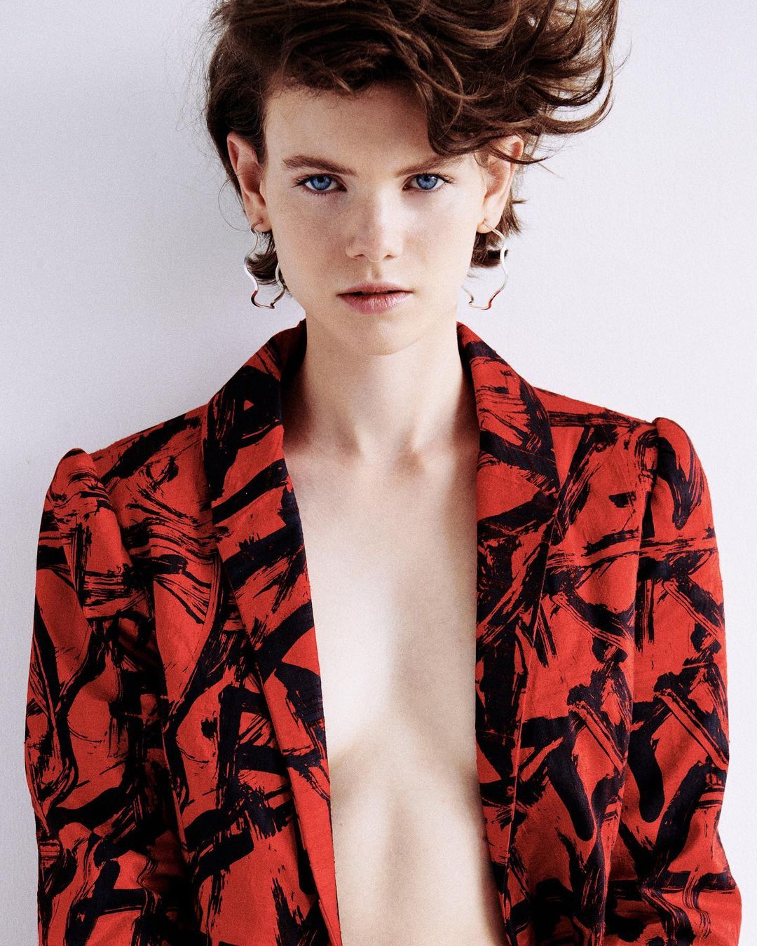 Portrait of a fair-skinned woman with short brown hair wearing a red and black patterned blazer against a white background.