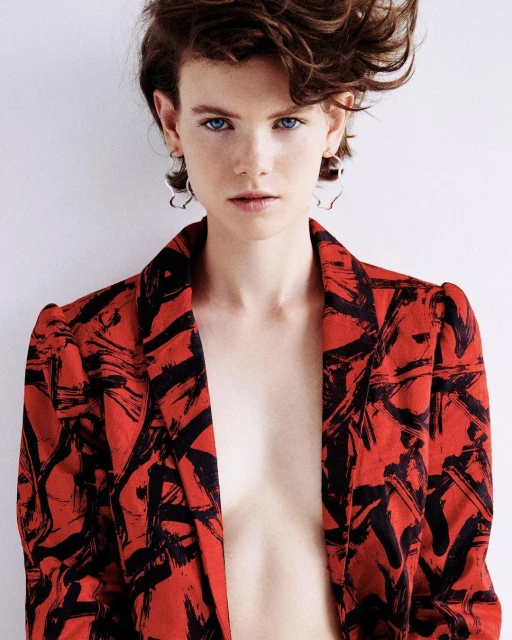 Portrait of a fair-skinned woman with short brown hair wearing a red and black patterned blazer against a white background.