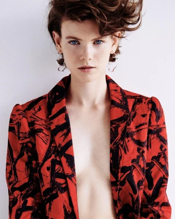 Portrait of a fair-skinned woman with short brown hair wearing a red and black patterned blazer against a white background.