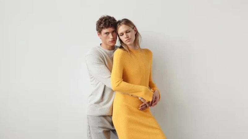 Man in a grey sweater embracing a woman in a yellow ribbed dress against a plain white studio background.