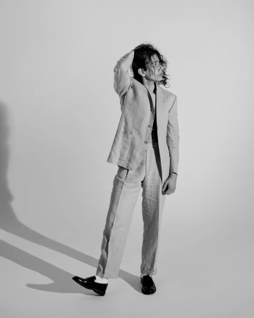 Portrait of a man with curly hair wearing a light-colored suit and dark loafers against a white backdrop.