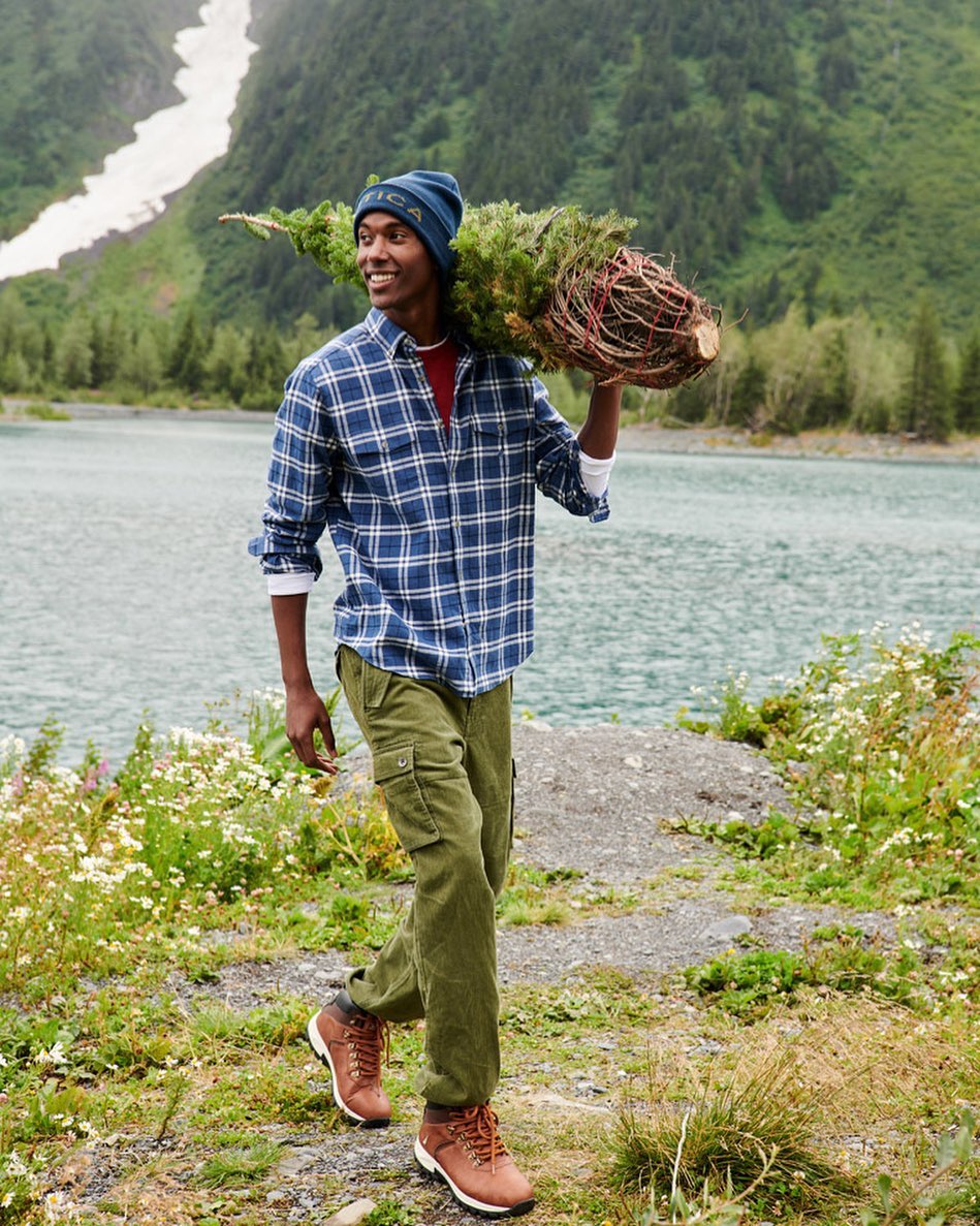 A smiling man carries a freshly cut Christmas tree over his shoulder near a lake in a lush mountain landscape, wearing a flannel shirt and beanie.