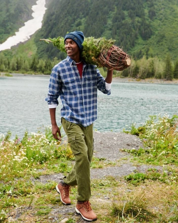 A smiling man carries a freshly cut Christmas tree over his shoulder near a lake in a lush mountain landscape, wearing a flannel shirt and beanie.