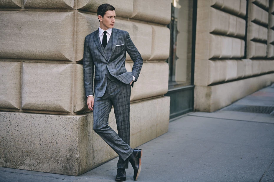 Man in grey plaid suit leaning against stone building, wearing black tie and dress shoes. Urban fashion shot, daylight, serious look.