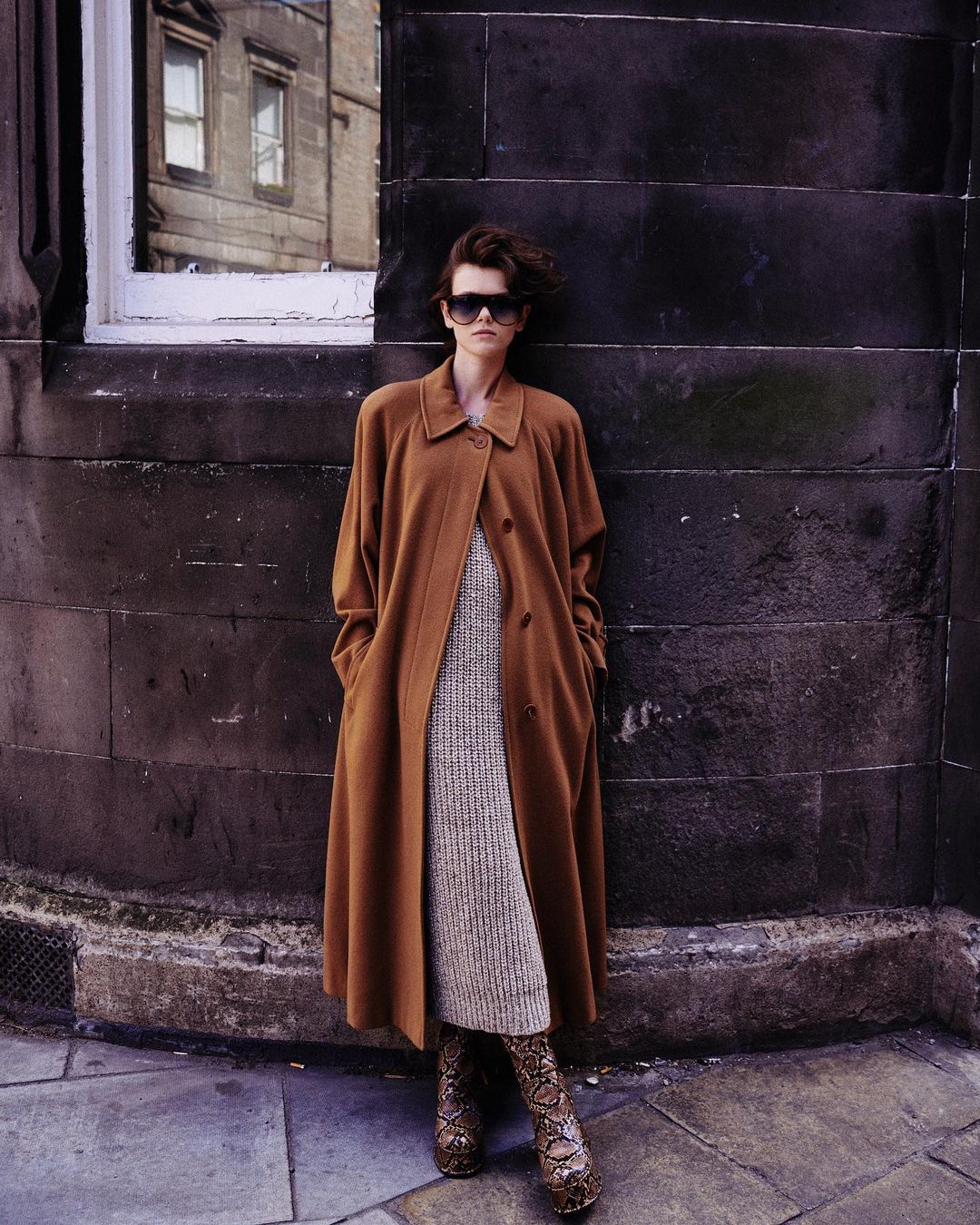 A stylish woman in a brown coat and snake print boots stands against a stone wall with a window, blending classic and bold fashion.