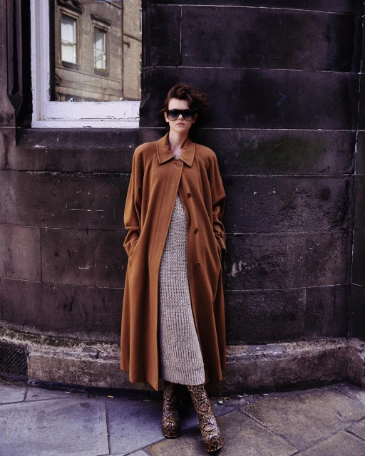 A stylish woman in a brown coat and snake print boots stands against a stone wall with a window, blending classic and bold fashion.