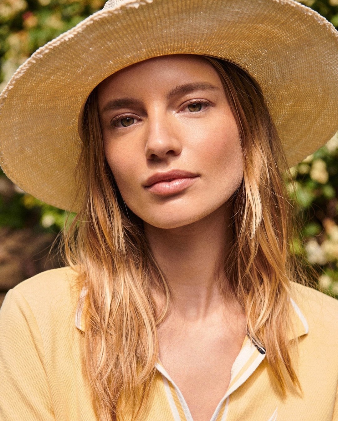 Close-up of a blonde woman wearing a woven sun hat and yellow collared shirt in an outdoor setting with green foliage backdrop.