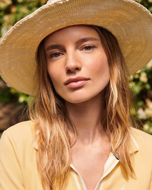 Close-up of a blonde woman wearing a woven sun hat and yellow collared shirt in an outdoor setting with green foliage backdrop.