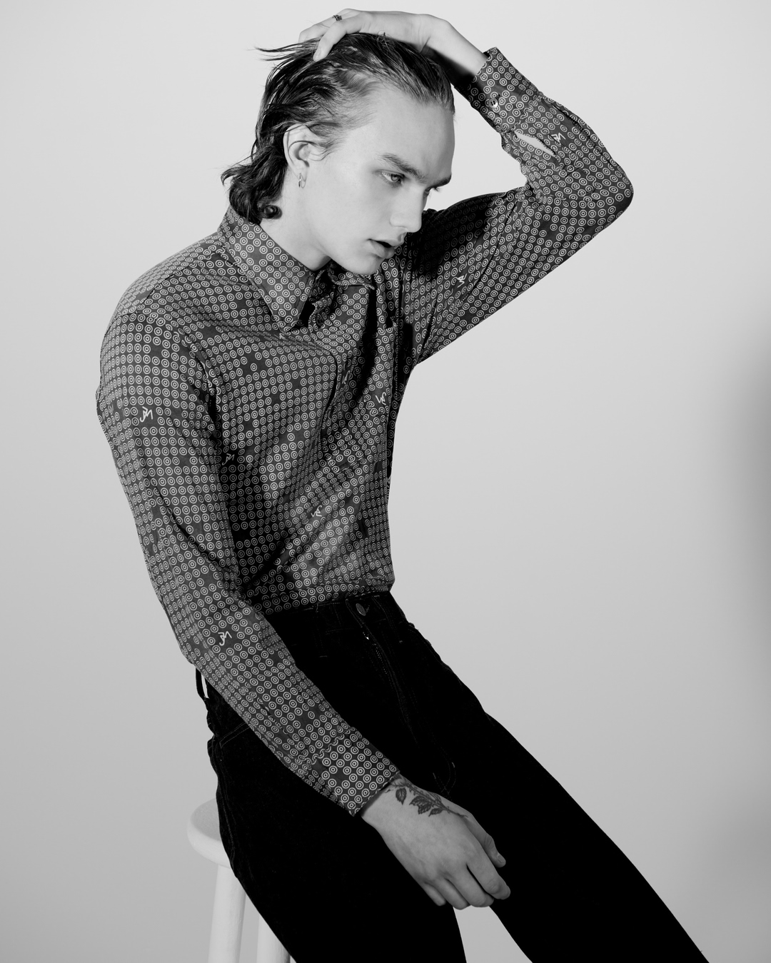 Black and white portrait of a young adult male model with dark hair slicked back, wearing a patterned shirt, seated on a white stool.