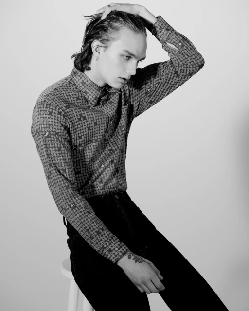 Black and white portrait of a young adult male model with dark hair slicked back, wearing a patterned shirt, seated on a white stool.