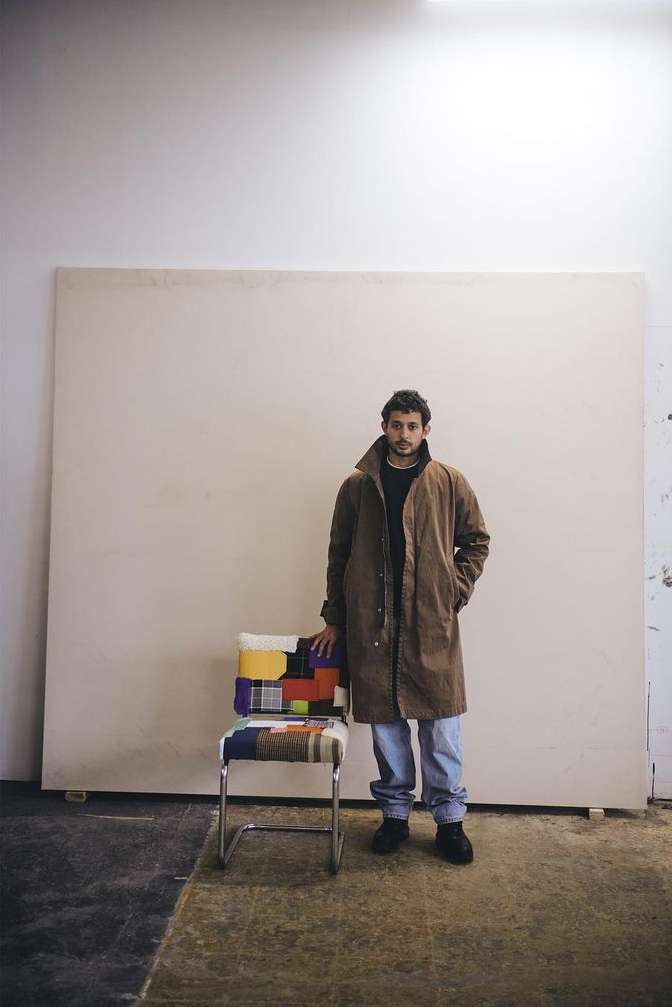 A man in a brown coat stands beside a colorful chair against a cream backdrop. He wears jeans and black shoes in a simple setting.