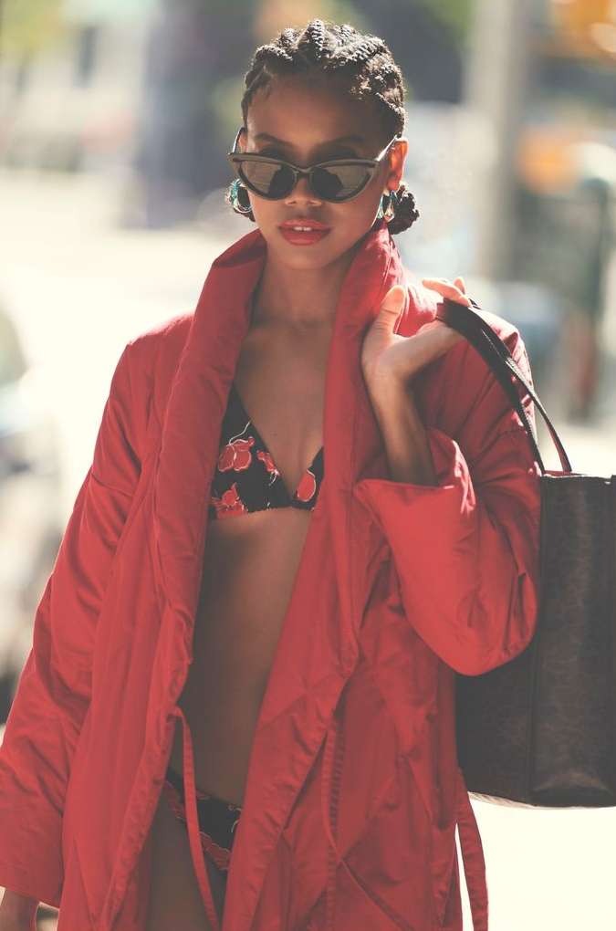 Woman in red coat and cherry-print bikini top and bottom stands outdoors holding a black tote bag and wearing sunglasses.