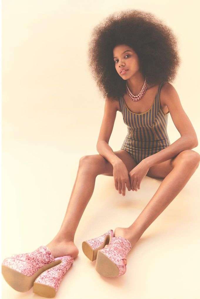 A person with dark skin and an afro sits indoors, wearing a swimsuit and pink platform shoes in a fashion-forward, beauty-focused shot.
