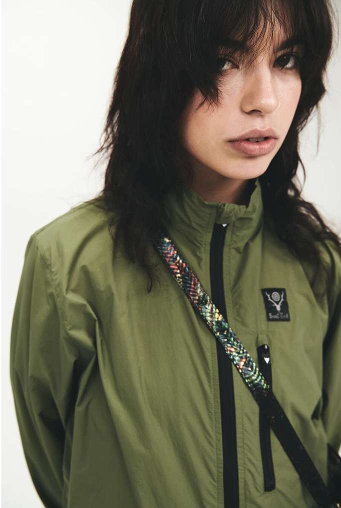 Close-up of a woman in an olive green jacket, featuring a braided crossbody strap and dark hair framing her face against a plain background.