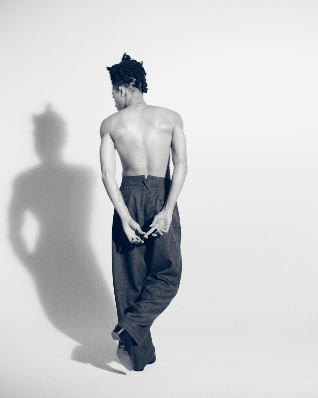 Shirtless man with braided hair wearing wide-leg trousers standing against a white seamless background with shadow