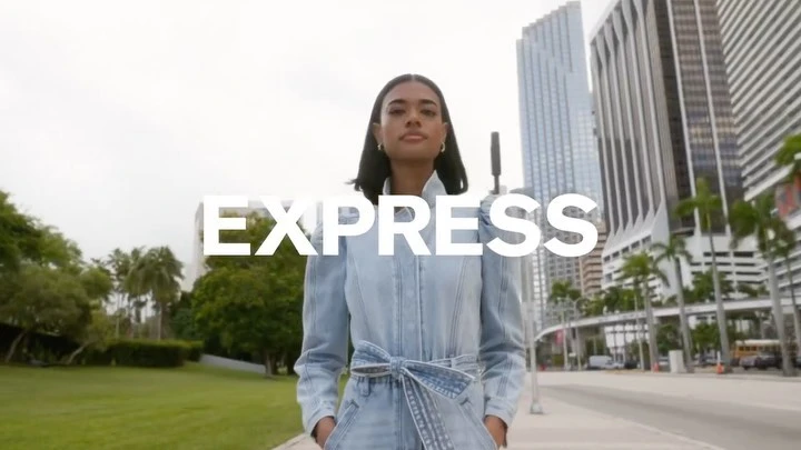 A woman in a denim jumpsuit walks toward the camera against a backdrop of city buildings and green trees, with the text EXPRESS.