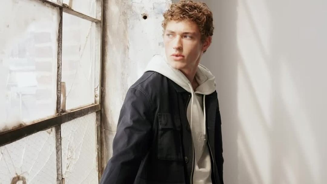 Young man with curly brown hair wearing a white hoodie and dark jacket near a large multi-pane window.