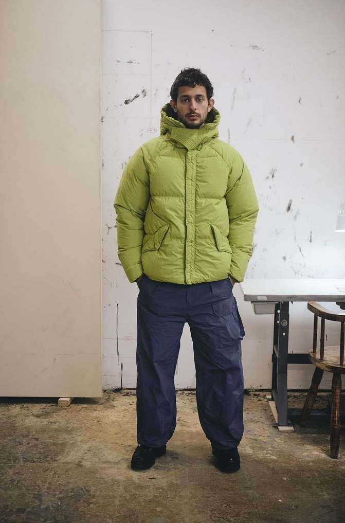 Adult male poses indoors wearing a lime green puffer jacket and blue pants, standing in front of a dirty white wall and furniture.