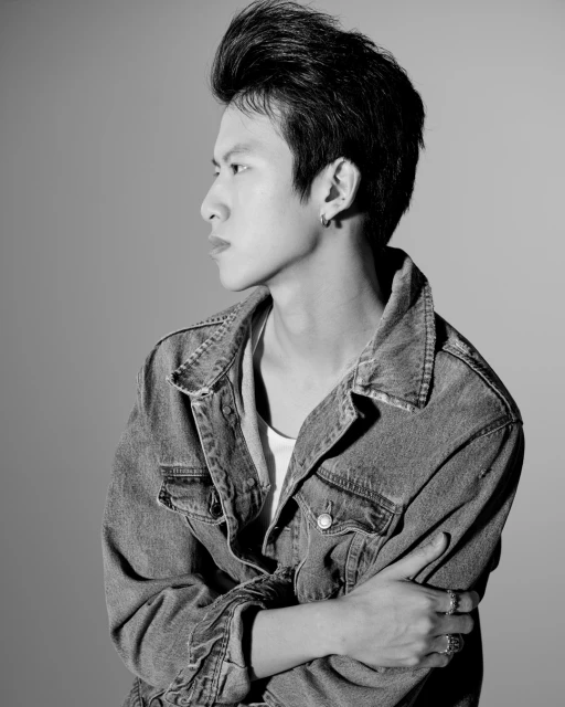 Monochrome portrait of an Asian man with pompadour, wearing a denim jacket, shot against a simple studio backdrop with even lighting.