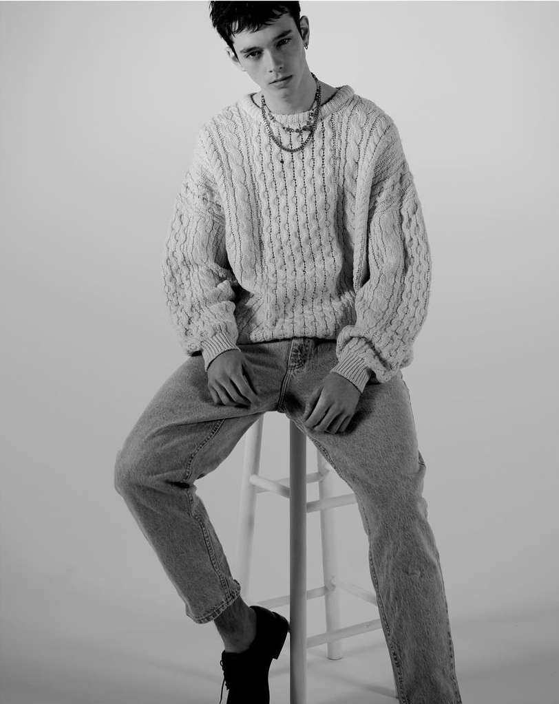 Monochrome portrait of a young man seated on a stool, wearing a cable knit sweater, light wash jeans, and black shoes, in a studio setting.