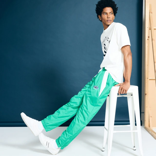 Young adult Black male in white logo t-shirt and green track pants sitting on white stool in a fashion studio against blue wall.