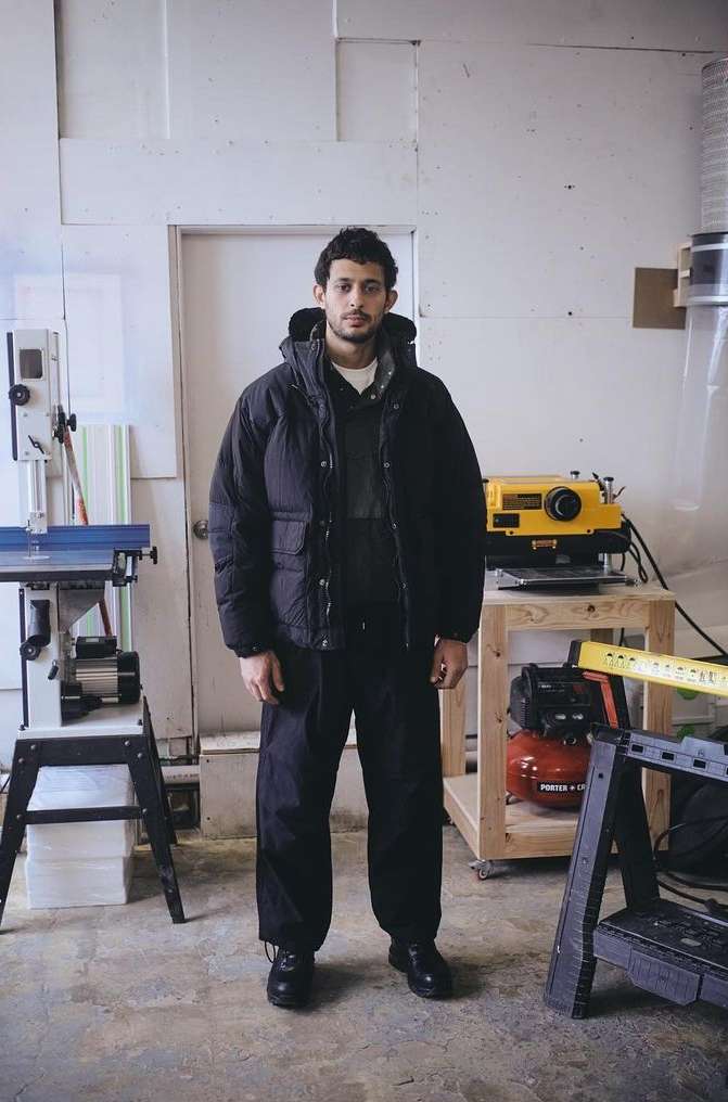 A man in a black puffer jacket stands in a workshop with tools and equipment visible in the background, looking toward the camera.