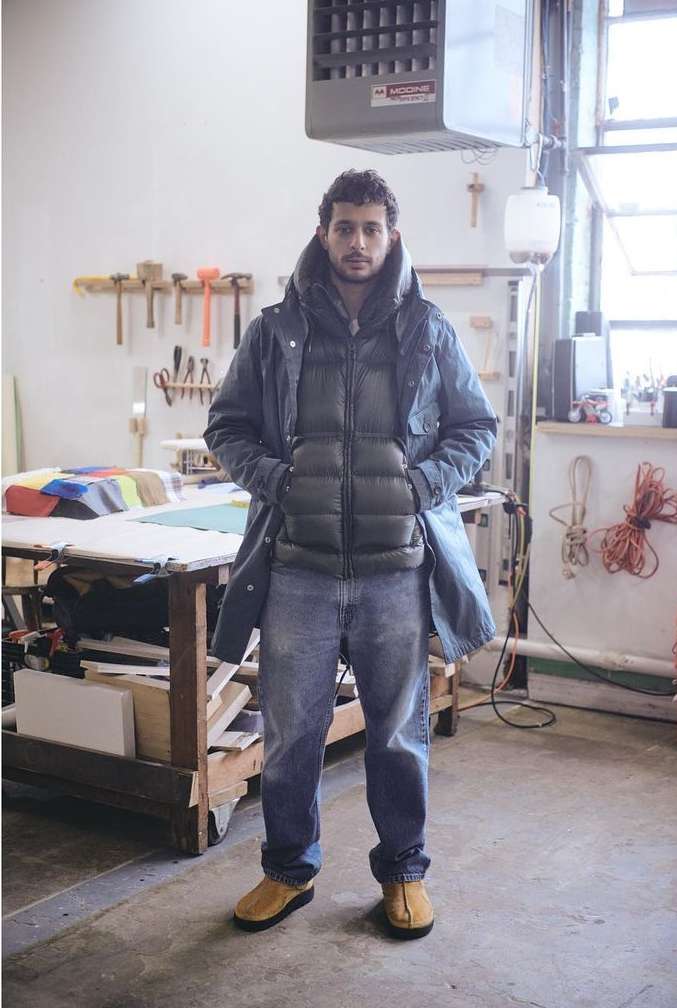Man in jacket and jeans poses indoors against background with workbench and tools; casual fashion, relaxed pose, neutral expression.
