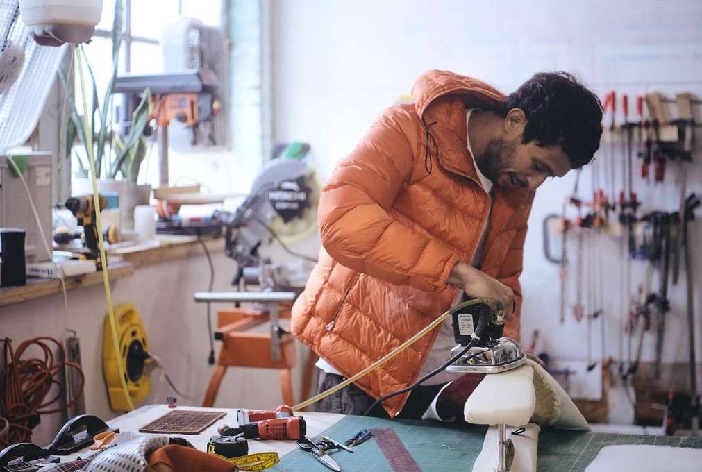 Adult man in orange puffer jacket working at a workbench with tools, sewing materials, and an iron in an indoor workshop setting.