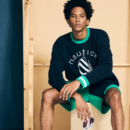 Black man seated wearing a navy blue sweatshirt with green trim and matching green shorts against a wooden set backdrop.