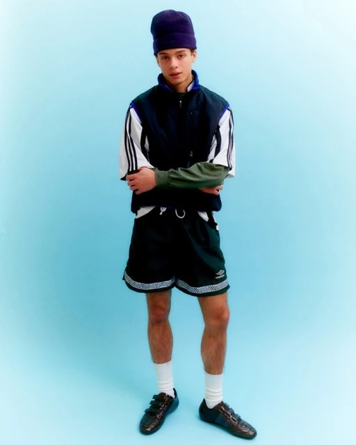 Young man in a sporty outfit stands against a blue background, wearing a navy vest, black shorts, socks, and shiny black shoes.