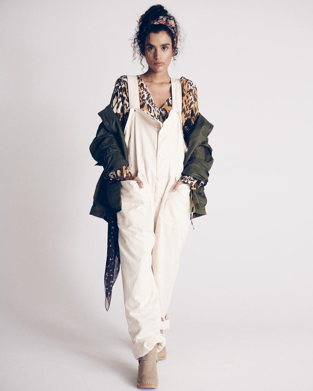 Full-shot of a woman in white overalls, an animal-print top, and a green jacket, standing in a photo studio with a white backdrop.